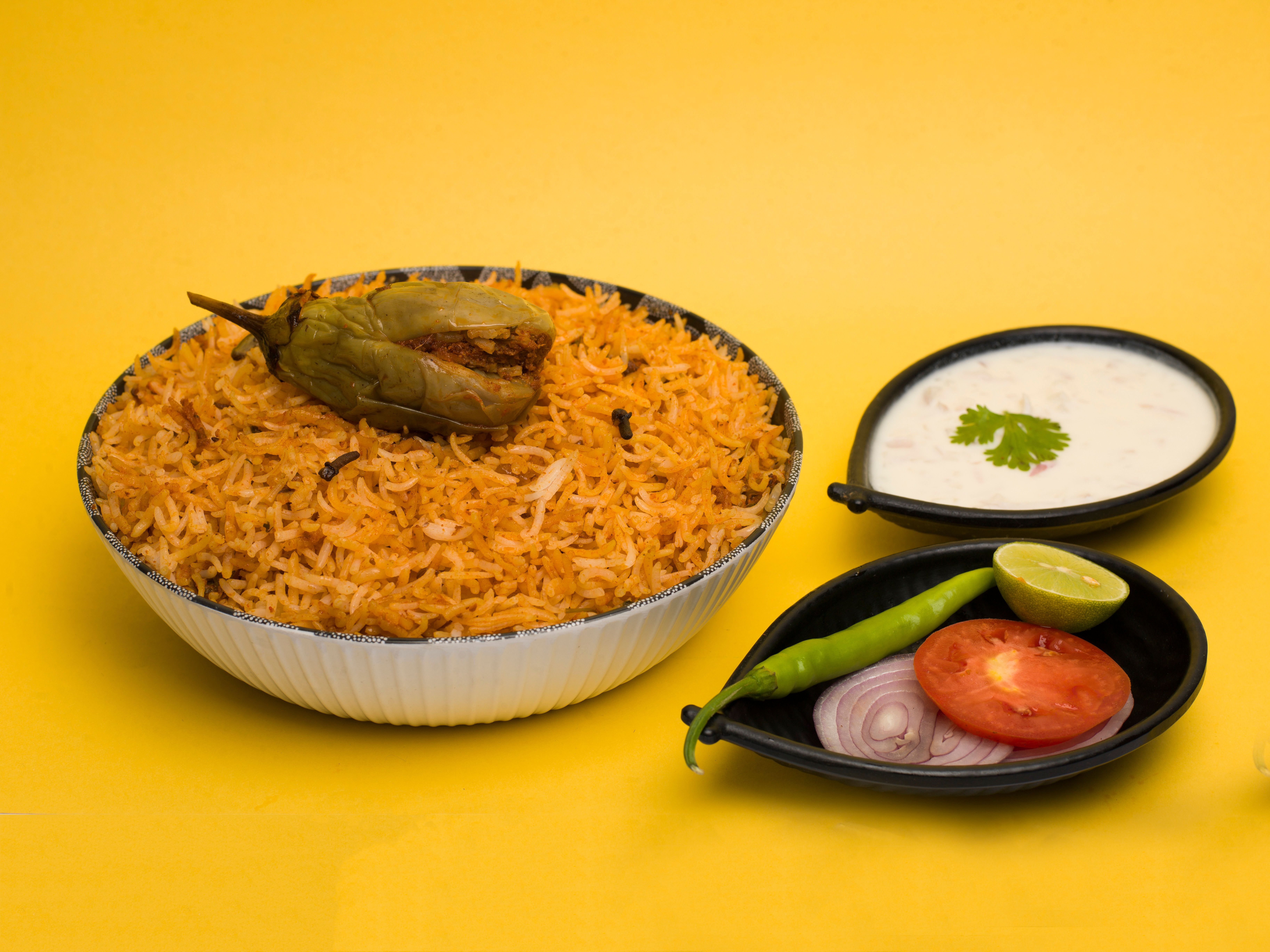 Spicy Indian biryani served with raita, chili, and vegetables on a vibrant yellow background.