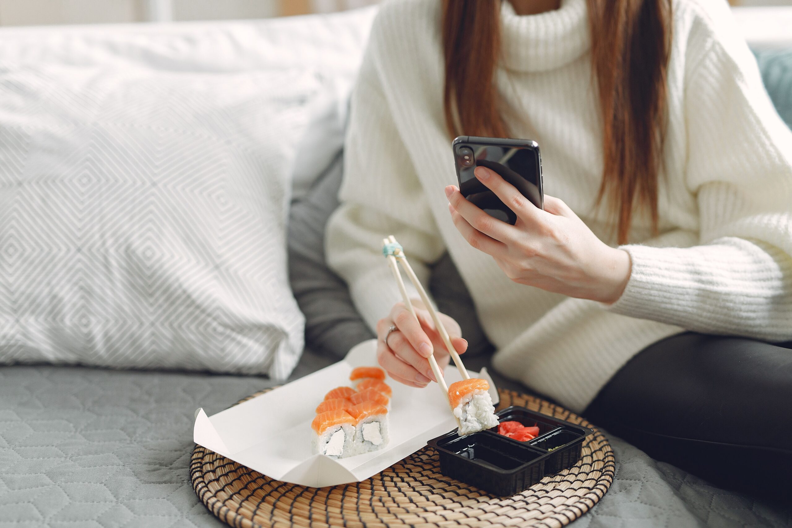 From above of unrecognizable woman in knitted sweater eating sushi with chopsticks and taking photo for social media whi